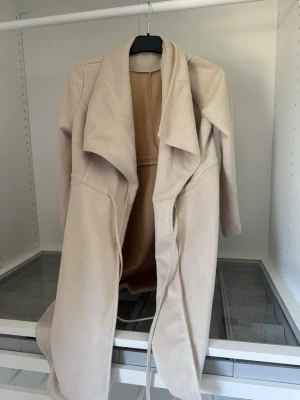 Beige lång kappa med skärp - Snygg beige kappa med lång passform och knytband i midjan. Kappan har en öppen front med breda slag och långa ärmar. Tillverkad i ett mjukt tyg som ger en stilren och modern look. Perfekt för dig som vill ha en enkel men trendig ytterjacka.