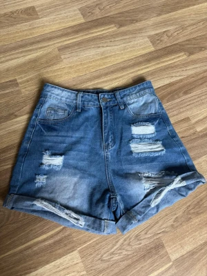 Blåa högmidjade jeansshorts - Snygga blåa jeansshorts med hög midja och slitna detaljer framtill och baktill. Shortsen har upprullade benslut, klassiska fickor och bälteshällor. Perfekta för en avslappnad och trendig look i sommar.