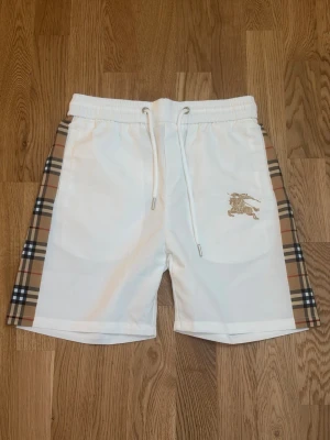 Shorts Burberry Check Panel Equestrian Knight Logo  - These white Burberry shorts feature classic check side panels and a prominent gold Equestrian Knight logo on the left thigh. Designed with an elastic drawstring waistband and side pockets, they offer a comfortable and stylish fit.  