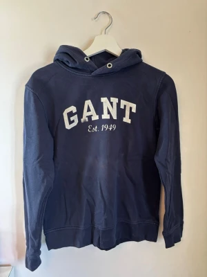 Mörkblå hoodie från GANT - Snygg mörkblå hoodie från GANT med vit broderad logga och texten 'Est. 1949' på bröstet. Perfekt för en avslappnad look. Storlek 158/164
