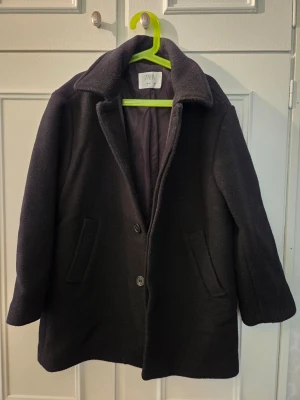 Svart kappa från ZARA 140 - Stilren svart kappa från ZARA med klassisk krage och två snedställda fickor framtill. Kappan har knäppning med knappar som döljs och är helfodrad för extra komfort. Perfekt för kyliga dagar och enkel att matcha med olika outfits. Finns lite ytludd/noppor. 