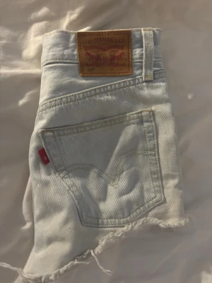 Levi's 501 ljusblå jeansshorts - Säljer ett par klassiska Levi's 501 jeansshorts i ljusblå denim. De har råa, fransiga benslut och den ikoniska läderpatchen bak. Perfekta för en avslappnad stil och har en snygg vintage vibe.
