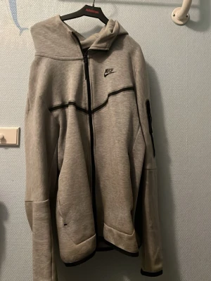 Grå Nike hoodie med dragkedja - Snygg grå hoodie från Nike med svart dragkedja och svarta detaljer. Klassisk logga på bröstet och huva för extra stilpoäng. Tillverkad i mjukt material som passar perfekt till chill eller streetwear. Fickor på sidorna och ribbade muddar vid ärmslut.