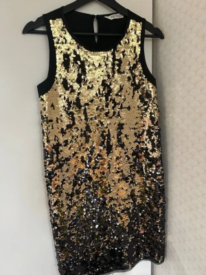 Svart och guld paljettklänning H&M - Snygg  klänning från H&M täckt med glittriga guld- och svarta paljetter. Ärmlös modell med rund halsringning och svart baksida. Perfekt för dig som vill sticka ut med en festlig look.