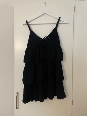 Svart volangklänning från Zara - Säljer en svart kort klänning från Zara med tunna axelband och volangdetaljer. Klänningen har omlottkänsla upptill med knyt framtill och är tillverkad i ett lätt, luftigt material. Perfekt för varma dagar eller sommarens alla tillfällen.