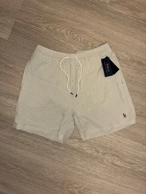 Randiga shorts från Polo Ralph Lauren - Snygga randiga shorts från Polo Ralph Lauren i vitt och beige. Shortsen har snörning i midjan och den klassiska broderade loggan på benet. Perfekta för sommardagar och har en avslappnad passform.