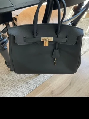 Svart klassisk handväska  - Stilren svart handväska i kraftigt material med två topphandtag och guldfärgat lås med hängande nyckel. Väskan har en strukturerad form och minimalistisk design, perfekt för dig som gillar tidlösa accessoarer. Passar till det mesta och rymmer allt du behöver.