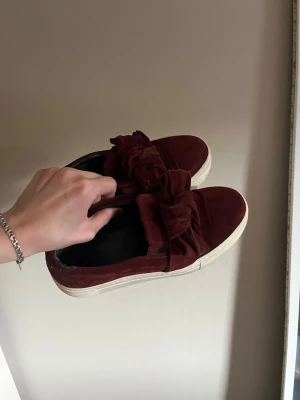 Vinröda sneakers med rosett - Snygga vinröda sneakers i mockaimitation med stora dekorativa rosetter på ovansidan och vit sula. Skorna har en slip-on design och låg profil som ger en clean look. Perfekta för dig som vill sticka ut med detaljer.