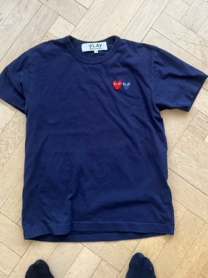 Mörkblå Comme des Garçons PLAY t-shirt - Säljer en mörkblå t-shirt från Comme des Garçons PLAY i bomull. T-shirten har korta ärmar och två broderade hjärtan i rött och blått på bröstet. Perfekt för dig som gillar stilrena plagg med en twist.