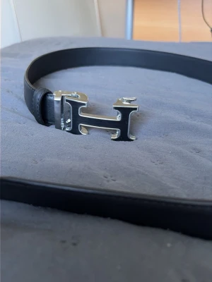 Svart bälte med silverspänne H - Snyggt svart bälte i skinn med ett stort H-format silverspänne. Perfekt accessoar för att lyfta din outfit och ge en stilren touch. Bältet har en klassisk design och passar till många olika looks.