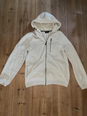 Beige Hoodie från Sail Racing - Säljer en stilren beige hoodie i nyskick med dragkedja framtill och en bröstficka med dragkedja. Använd fåtal gånger men säljs pågrund av bara hängande i garderob!Perfekt för en avslappnad look och enkel att matcha med andra plagg.    Nypris:1500kr Mitt pris:550kr