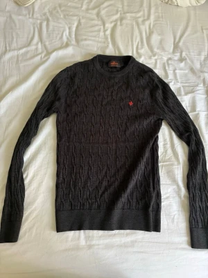 Morris tröja - Snygg mörkgrå kabelstickad tröja med rund halsringning och långa ärmar. Tröjan har ett diskret rött broderat emblem på bröstet och ribbade muddar vid ärmslut och nederkant. Perfekt för lager på lager och en stilren look.