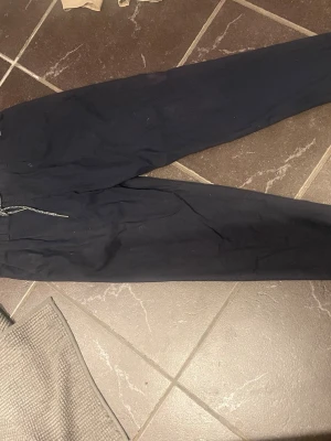Mörkblå mjukisbyxor med dragsko - Säljer ett par mörkblå mjukisbyxor med dragsko i midjan och raka ben. Byxorna har en enkel och clean look, perfekta för chill dagar. Materialet känns mjukt och bekvämt mot huden.