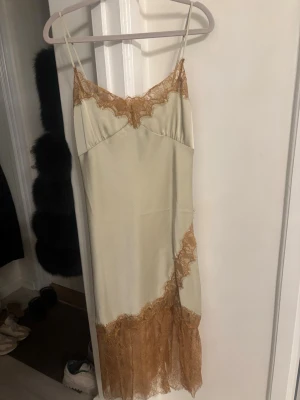 Mintgrön  långklänning med spets - Säljer en elegant långklänning i champagnefärg med tunna axelband och dekorativa spetsdetaljer i brunt vid byst och nederkant. Klänningen har en mjuk, glansig yta och en feminin siluett som ger en lyxig känsla.