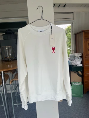 Vit sweatshirt från Ami Paris - Snygg vit sweatshirt från Ami Paris med röd hjärtlogga på bröstet. Tröjan har rund halsringning, långa ärmar och ribbade muddar. Perfekt för en clean och stilren look. Materialet är mjukt och bekvämt, passar till många outfits.