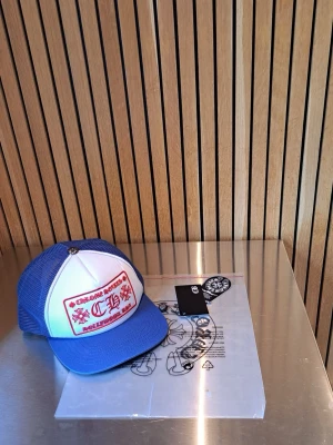 Blå och vit truckerkeps Chrome Hearts - Snygg truckerkeps från Chrome Hearts i blått och vitt med röd broderad logga framtill. Kepsen har mesh på sidorna och bak, samt en metallknapp på toppen med märkets ikoniska detalj. Justerbar passform och böjd skärm.
