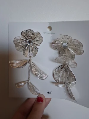 Silvriga blommiga örhängen H&M - Unika silvriga örhängen från H&M i form av stora blommor med transparent sten i mitten. Örhängena har hängande blad och detaljer i metalltråd som ger en luftig och modern look. Perfekta för dig som vill sticka ut med accessoarerna.