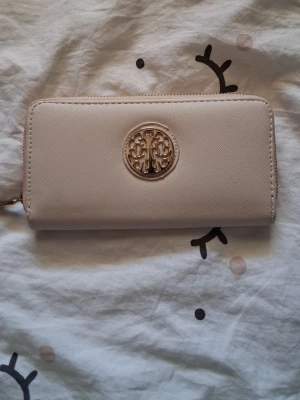 Beige plånbok med gulddetalj - Stilren beige plånbok i konstläder med guldfärgad dragkedja och ett dekorativt guldfärgat emblem på framsidan. Plånboken har en rektangulär form och stängs med dragkedja runt om. Perfekt för dig som gillar enkel och elegant design.