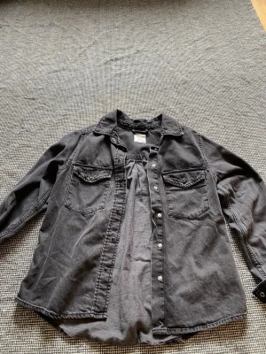 Svart overshirt i denim - Säljer en svart overshirt i denim med tryckknappar framtill och på ärmsluten. Två bröstfickor med lock och klassisk krage. Oversized så jag som vanligtvis har M i stjortor kan ha den här öppen över en tshirt. Använd en gång 