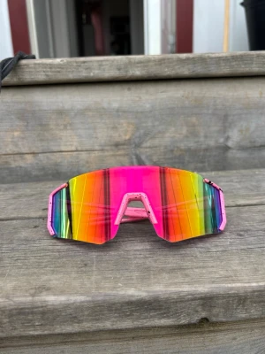 Rosa Sportglasögon Färgglada sportiga solglasögon - Unisex. One size. Säljer ett par riktigt färgstarka solglasögon med spegelglas i regnbågens färger och rosa bågar. Glasen har en sportig, kantig form och coola ränder som ger en unik look. Perfekta för dig som vill sticka ut i sommar och vår!