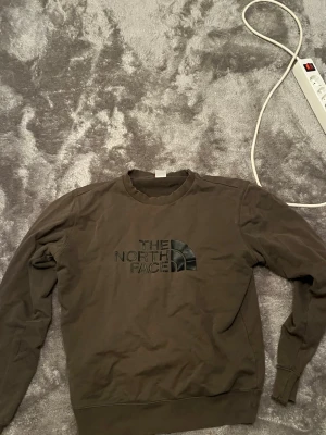 Mörkgrön sweatshirt från The North Face - Säljer en mörkgrön sweatshirt från The North Face med stor logga i svart på bröstet. Tröjan har rund halsringning, långa ärmar och ribbade muddar vid ärmslut och nederkant. Perfekt för en avslappnad och stilren look.