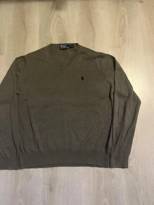Polo Ralph Lauren Tröja – Storlek L - Säljer en riktigt snygg och stilren stickad tröja från Polo by Ralph Lauren i storlek L Tröjan är i en fin grå  melerad nyans med broderad mörk hästlogga på bröstet. Tillverkad i en mjuk och skön kvalitet med resårstickning i ärmslut och midja, samt helt slät baksida. Perfekt att bära över en skjorta eller som den är. Tröjan är i mycket fint begagnat skick utan skador eller fläckar. 