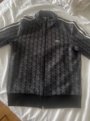 Svart mönstrad hoodie från Lacoste - Snygg svart hoodie från Lacoste med hel dragkedja och diskret mönster över hela plagget. Har vita ränder på axlarna och den klassiska Lacoste-loggan på bröstet. Ribbstickade muddar vid ärmslut och nederkant. Perfekt för en sportig och trendig look.