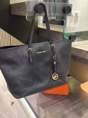 Svart toteväska från Michael Kors - Stilren svart toteväska från Michael Kors i strukturerat läder med guldfärgade detaljer och MK-hänge. Väskan har dubbla axelband och logotyp framtill. Perfekt för dig som vill ha plats för allt du behöver och samtidigt se trendig ut.
