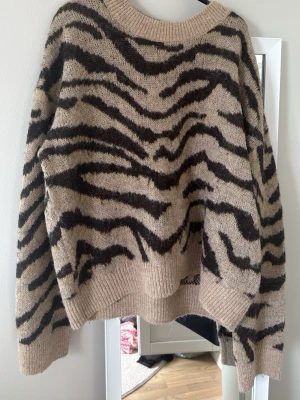 Beige stickad tröja med zebramönster - Stickad tröja i beige med brunt zebramönster. Normal passform med rund halsringning och ribbade muddar vid ärmslut och nederkant. Perfekt för dig som gillar djurmönstrat 