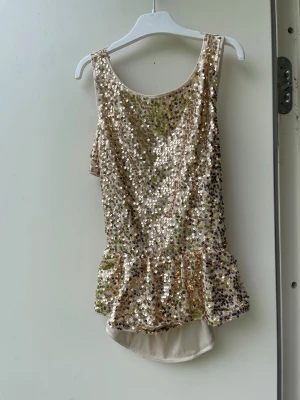 Guldig topp med paljetter och volangkant - Säljer en glittrig topp i guld med paljetter och volangkant nedtill. Toppen har ärmlös design och rund halsringning, perfekt för dig som vill sticka ut lite extra. Från Bik bok storlek S