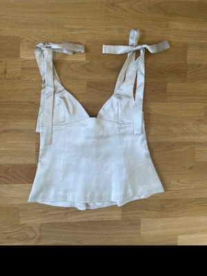 Vit linnetopp med knytband - Snygg vit linnetopp med breda axelband som knyts upptill. Toppen har en lätt utsvängd passform och är tillverkad i ett mjukt, lite glansigt material som ger en lyxig känsla. Perfekt att styla med jeans eller kjol för en fräsch look.