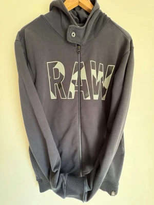 Zip-Hoodie med RAW-tryck - Snygg hoodie med dragkedja och stort RAW-tryck i vitt och grått på bröstet. Huvan har hög krage med knappdetalj och tröjan är gjord i mjukt bomullsmaterial. Perfekt för en avslappnad streetwear-look.