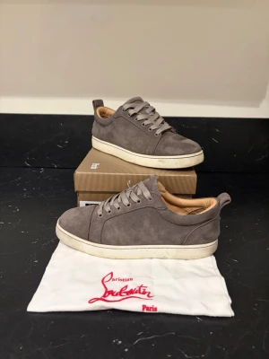 Louboutin - Snygga gråa sneakers i mocka från Christian Louboutin med klassisk röd sula och vita kanter. Skorna har rund tå, platta skosnören och beige innersida. Perfekta för dig som vill ha en stilren och lyxig look.