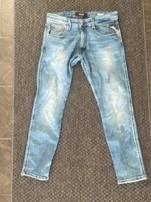 Ljusblå slim fit jeans replay - Säljer ett par ljusblå slim fit jeans med slitningar och tvättade detaljer. Klassisk femficksmodell med dragkedja och knapp i midjan. Snygga sömmar på bakfickorna och smal passform som sitter tajt längs benen. Perfekta till en avslappnad streetstil.
