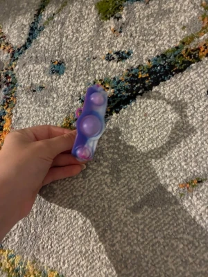 Fidget pop armband i lila och rosa - Säljer ett färgglatt fidget pop armband i mjukt silikonmaterial. Armbandet har en mix av lila, blå och rosa nyanser samt tre poppbara bubblor som är perfekta att pilla på när man har tråkigt eller vill slappna av. Justerbar storlek och passar de flesta handleder.