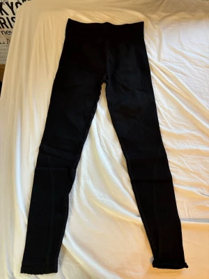 Svarta ribbade leggings med hög midja - Säljer ett par svarta leggings med hög midja och ribbade detaljer längs benen. De har en tight passform och är stretchiga, perfekta för en trendig och bekväm look. Materialet känns mjukt och följsamt, vilket gör dem sköna att bära hela dagen.