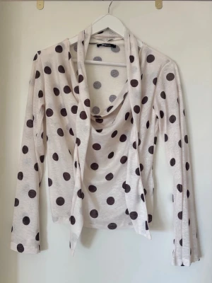 Prickig långärmad topp med scarfdetalj - Säljer en vit långärmad topp med stora svarta prickar och en snygg scarfdetalj vid halsen. Toppen har en lös passform och är tillverkad i ett mjukt, tunt material som känns skönt mot huden. Perfekt för dig som gillar lekfulla mönster och trendiga detaljer. Från Gina! Aldrig använd!