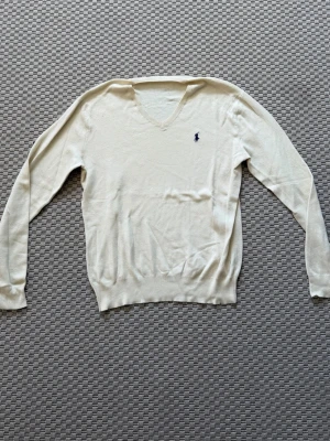  stickad tröja från Ralph lauren - Klassisk beige stickad tröja från Ralph Lauren med V-ringning och diskret mörkblå logga broderad på bröstet. Långärmad modell i tunt, mjukt material som passar perfekt till lager på lager. Ribbstickade muddar vid ärmslut och nederkant. Tvivla inte att skriva vid funderingar!