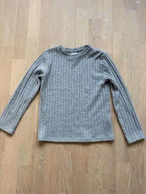 Grå ribbstickad tröja med pärlor - Snygg grå ribbstickad tröja med långa ärmar och rund hals. Tröjan har dekorativa små pärlor framtill som ger en extra touch. Perfekt till jeans eller kjol för en trendig look.