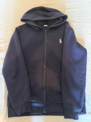 Mörkblå hoodie med dragkedja - Snygg mörkblå hoodie med dragkedja och huva. Klassisk design med två fickor framtill och en broderad logga på bröstet. Perfekt för en avslappnad stil och enkel att matcha med jeans eller joggers.