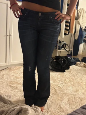 Vintage mörkblå bootcut jeans  - Säljer ett par mörkblå bootcut jeans med lätt slitna detaljer och klassiska fem fickor. Jeansen har låg midja och stängs med knapp och dragkedja. Perfekta för en avslappnad och trendig look.