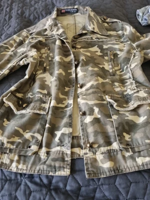 Kamo vindjacka med knappar  - Snygg vindjacka i camouflage-mönster från River Flower.Jackan har flera fickor med dragkedjor och knappar, samt detaljer vid ärmslut och axlar. Tillverkad i ett lätt tyg som passar perfekt för blåsiga dagar. Klassisk krage och avslappnad passform. I väldigt fin skick. Storlek är inte säkert men kan passa M. 