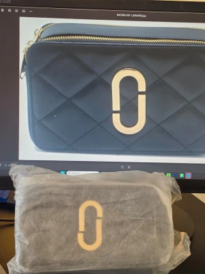 Svart quiltad handväska med gulddetaljer - Snygg svart quiltad handväska med två guldfärgade dragkedjor och en stor guldfärgad logga framtill. Väskan har en bred axelrem med svartvitt grafiskt mönster och rektangulär form. Perfekt för dig som vill ha en trendig accessoar med plats för det viktigaste.