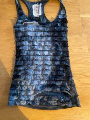 Svart volang-linnetopp - Säljer en svart linnetopp med volangdetaljer i flera lager framtill. Toppen är ärmlös och har en racerback-design. Materialet känns lätt och toppen har en snygg glansig finish som ger extra edge till din outfit.