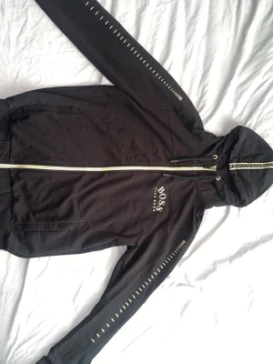 Hugo Boss hoodie - Snygg svart hoodie från BOSS med vit dragkedja och logga på bröstet. Hoodien har huva med snörning och dekorativa vita detaljer längs ärmarna. Perfekt för en avslappnad och trendig stil. Storlek M passar mig på 186cm och 78kg