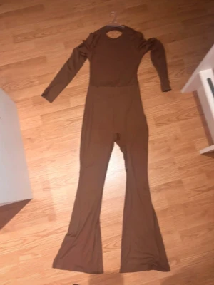 Brun långärmad jumpsuit - Säljer en brun långärmad jumpsuit med figurnära passform och utsvängda ben. Materialet ser stretchigt och mjukt ut, perfekt för dig som gillar en enkel men trendig look. Rund halsringning och hel längd på benen.