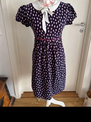 Mörkblå klänning med vit krage Mademoiselle R - Söt mörkblå klänning med små blommiga mönster i vitt och rosa. Klänningen har en stor vit krage med knytband, puffärmar och markerad midja med rosa kant. Perfekt för dig som gillar vintageinspirerad stil. Storlek 34, jättefint skick, märke Mademoiselle R