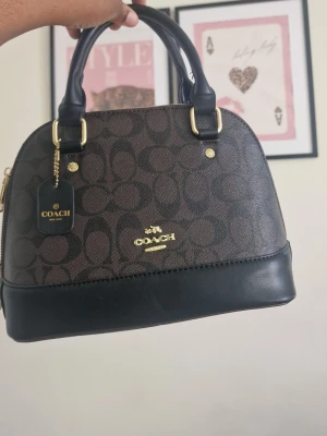 Brun och svart handväska - Snygg handväska i brunt och svart konstläder med klassiskt logomönster. Väskan har guldfärgade detaljer, dubbla handtag och en stabil botten med metallfötter. Perfekt för dig som gillar stilrena accessoarer med lyxig känsla.