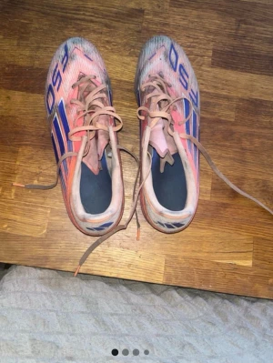 Orange Adidas F50 fotbollsskor - Säljer ett par rosa Adidas F50 fotbollsskor med blå detaljer och snörning. Skorna har en låg profil och är designade för fart på planen. Materialet är syntetiskt och skon har en smal passform som ger bra bollkontroll.