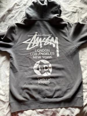 Grå Stüssy x i-D hoodie - Säljer en grå hoodie från Stüssy i samarbete med i-D. Stor vit logga på ryggen med text om London, Los Angeles, New York, Tokyo och Paris. Tryck även på framsidan med i-D och krona. Klassisk huva och avslappnad passform. Perfekt för streetwear-stilen.